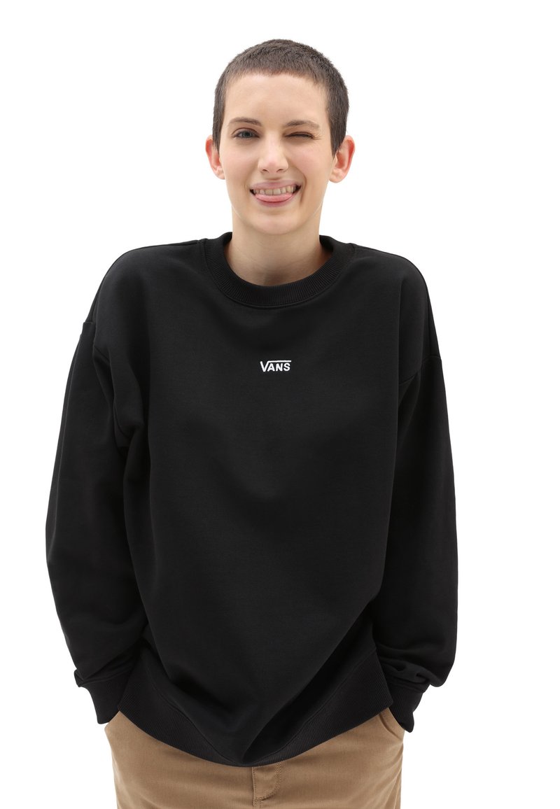 Zwarte sweatshirt met een ronde hals en lange mouwen, voorzien van een wit "VANS" logo op de borst. Gedragen over beige broek.
