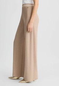 Pantaloni beige a gamba larga realizzati in un tessuto morbido e strutturato. Presentano una cintura a righe in contrasto e dettagli in metallo dorato.