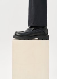 Sorte skinnloafers med et strømlinjeformet design, med solid gummisåle og sydd detaljer, vist på en treblokk.