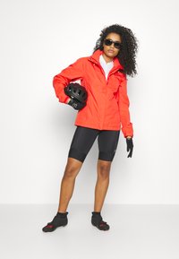Red waterproof jacket over a white shirt, black cycling shorts, gloves, and sunglasses. Model holding a black bicycle helmet. Smooth textures.