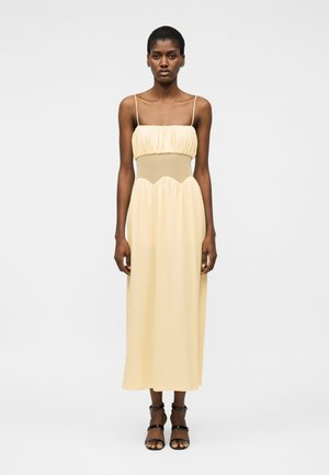 Γυναίκα που στέκεται, φοράει ένα χωρίς μανίκια πλάτινι κίτρινο μίντι φόρεμα με λεπτές τιράντες και μαύρα ανοιχτά τακούνια, μπροστά σε σκηνικό λευκού φόντου.