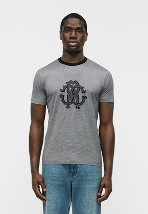 Homme portant un t-shirt gris à manches courtes avec un col noir et un monogramme noir orné, associé à un jean bleu.