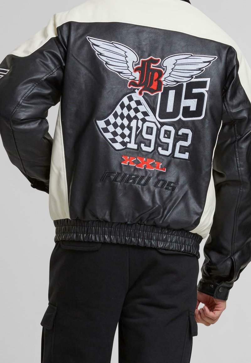 Veste bomber noire et crème avec logo ailé, motif à carreaux et accents rouges. Comprend une ceinture élastique et des poignets côtelés. Taille XXL.