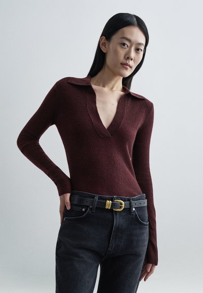 Bordeauxrød ribstrikket top med dyb V-hals og krave, kombineret med højtaljede sorte jeans og et sort bælte med en guldbeklædt spænde.