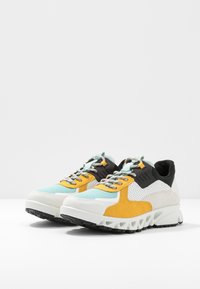 Sport sneakers som har en blandning av mintgrön, svart, vit och gul material med nätakcenter och texturerade skosnören på en gummisula.