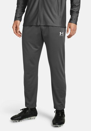 Mann mit dunkelgrauer Under Armour Trainingshose und schwarzen Fußballschuhen steht vor einem einfarbigen Hintergrund.