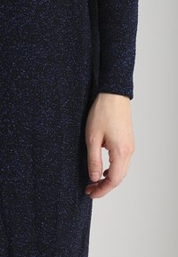 Schwarzes Kleid aus strukturiertem Stoff mit einem blauen Schimmer. Die Ärmel sind lang, und eine Hand ruht auf dem Kleid, was die Passform zur Geltung bringt.