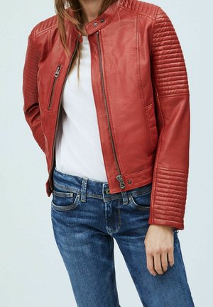 Femme portant une veste en cuir rouge avec des manches matelassées, une chemise blanche et un jean bleu, debout avec une main détendue le long du corps.