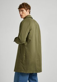 Olijfgroene trenchcoat met een kraag, lange mouwen, zijzakken en een rechte snit, gecombineerd met blauwe spijkerbroek.