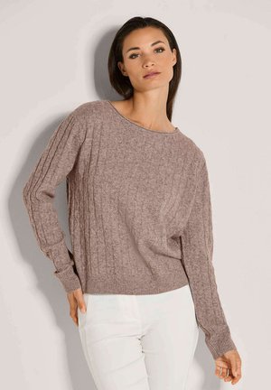 Frau in einem hellbraunen Kabelstrickpullover und weißen Hosen, die mit einer Hand auf der Hüfte vor einem schlichten hellen Hintergrund posiert.