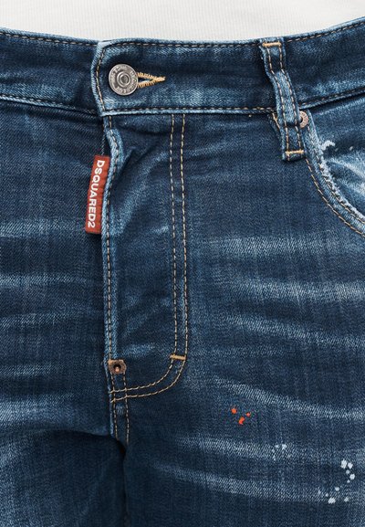 Primo piano di jeans in denim blu con cuciture arancioni, bottone di metallo, passante per cintura e etichetta rossa DSQUARED2 sul bordo della tasca.