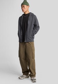Felpa grigia con zip sopra una maglietta nera, abbinata a pantaloni cargo color oliva e sneakers grigie. Include un berretto verde e collane sovrapposte.