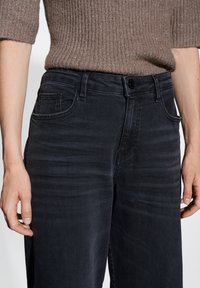 Jeans bleu foncé avec une texture lisse, design à cinq poches et fermeture à bouton. Le modèle porte un pull marron côtelé ajusté.