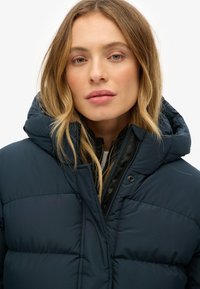 Giacca a piumino blu navy scuro con cappuccio; presenta una trama trapuntata e una fodera interna a contrasto. Chiusura con cerniera e bottoni a pressione visibili.