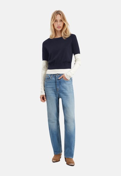 Maglione cropped bicolore navy e crema con maniche corte, abbinato a jeans a gamba dritta azzurri chiari e stivali alla caviglia marroni.