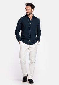 Marineblauw overhemd met knopen in textuurstof, slim fit, met witte knopen, gecombineerd met een witte broek en zwarte schoenen. Model staat casual.