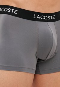Sive bokserice iz gladkega materiala, z črnim pasom, na katerem je belo napisano "LACOSTE". Prilagojen kroj z brezšivnimi stranmi.