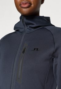 Chaqueta de cremallera azul marino con un alto cuello, textura suave y un bolsillo en el pecho visible con una cremallera negra y acento de logo.