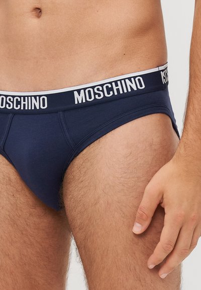 Tamsiai mėlynos medvilninės kelnaitės su baltais Moschino logotipo juostos elementais. Audinys yra lygus, o dizainas pasižymi prigludusia forma ir minimalistiniu stiliumi.