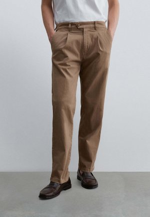 Hombre con pantalones plisados marrón claro, camisa blanca metida dentro y mocasines de cuero marrón oscuro, de pie con las manos en los bolsillos.