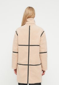 Cappotto in pile beige chiaro con collo alto, caratterizzato da cuciture a griglia nere. Il cappotto ha maniche lunghe e una vestibilità ampia.