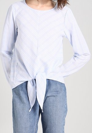 Hellblaues Langarmoberteil mit einem Chevron-Streifenmuster und einer gebundenen Vorderseite, kombiniert mit blauen Denim-Jeans.