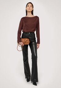 Haut long à manches en satin marron associé à un pantalon évasé en simili cuir noir. Le mannequin tient un petit sac à main marron avec une double sangle. Talons aiguilles noirs.