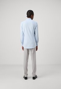 Cămășă albastră deschis cu mâneci lungi, hem curbat, manșete cu nasturi. Asortată cu pantaloni gri deschis și pantofi negri. Textură de țesătură netedă.