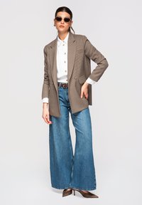 Femme portant un blazer à carreaux marron, une chemise blanche boutonnée, un jean à jambes larges bleu, des talons marron et des lunettes de soleil rondes, debout avec une main sur la hanche.