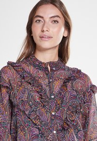 Blusa de manga larga con un estampado paisley oscuro en colores vibrantes. Presenta acentos de volantes a lo largo del escote y la parte frontal. Confeccionada en tejido transparente.