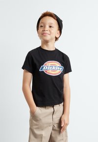 Juodas medvilninis marškinėlis su ryškiu ovaliu Dickies logotipu. Derinami su smėlinėmis kelnėmis ir juoda kepure. Paprastas ir kasdieniškas dizainas.