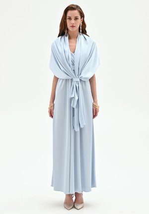 Donna in un vestito azzurro chiaro lungo fino a terra con uno scialle abbinato legato in vita, indossa orecchini d’oro, braccialetti e scarpe a punta.