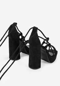 Sandalias de tacón alto de ante negro con tacón sólido, punta abierta y diseño de tiras entrelazadas, que cuentan con lazos largos para un ajuste seguro.