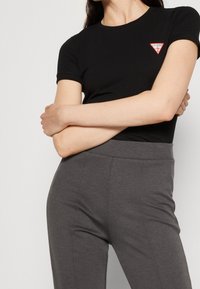 Černé tričko s kulatým výstřihem a červeným a bílým trojúhelníkovým logem na levé hrudi, doplněné šedými přiléhavými kalhotami. Tkanina vypadá hladce.