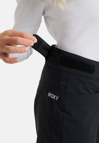 Mustat housut, joissa on tekstuurinen vyötärönauha ja tarranauhak closure, esittelee "ROXY"-logoa valkoisella brodeerauksella sivulla.