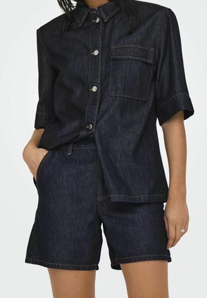 Femme portant une chemise à manches courtes en jean foncé avec poche poitrine et un short en jean taille haute assorti.