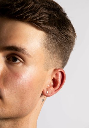 Sølv lynlås ørepine bæres i højre øre; kortklippet hår; synlig kors-tatovering på halsen; glat hudtekstur.