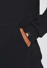 Sudadera negra con bolsillo canguro, puños acanalados y una pequeña etiqueta de logo en la manga con acentos en naranja y rojo. Textura suave.