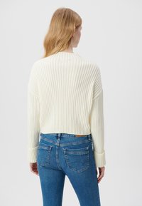 Gekürzter Strickpullover in Off-White mit gerippter Textur und weiten Ärmeln, kombiniert mit blauen, schmal geschnittenen Jeans mit Gesäßtaschen.