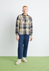 Rutigt flanellskjorta i beige och marinblå, uppställd med mörkblå jeans och beige sneakers. Mjuk textur med knäppt fram och fickor.