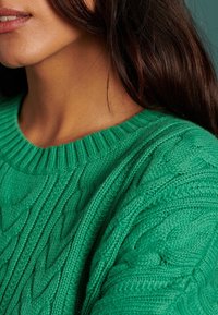 Suéter verde de punto con cuello acanalado y patrones de trenzas texturizadas. El primer plano enfatiza la rica y suave textura y el color vibrante.