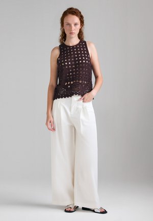 Jeune femme portant un haut noir en crochet sans manches, un pantalon blanc à jambes larges et des sandales noires et argentées, se tenant devant un fond gris uni.