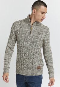 Gebreide trui in beige en bruine tinten, met een kwart ritssluiting, kabelgebreide textuur en een geweven merkembleem aan de onderzijde van de voorkant.