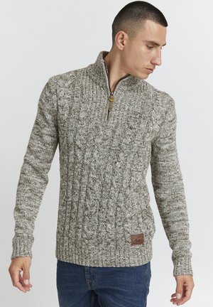 Knitted sweater in beige and brown tones, featuring a quarter zip, cable-knit texture, and a textured brand patch on the lower front.
