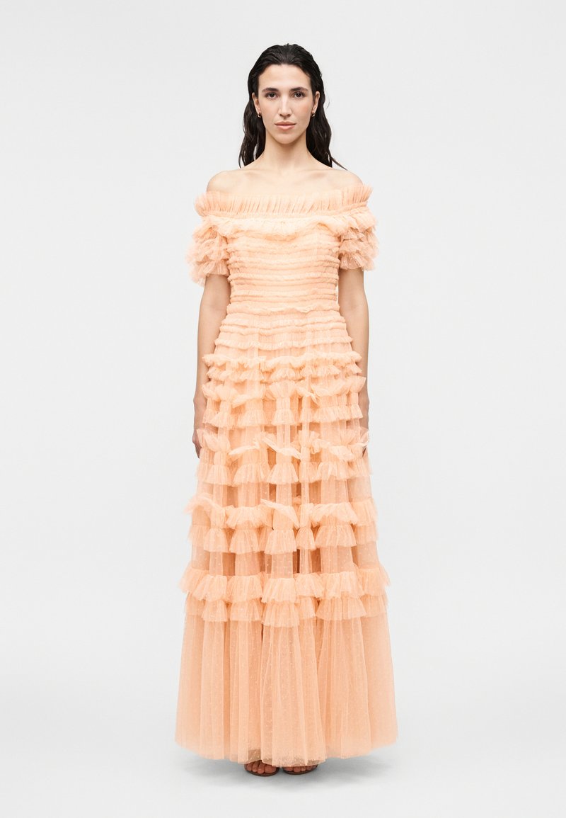 Naine kannab virsikuvärvi õlgadeta kleiti, millel on kihilised voliütlid ja läbipaistev kangas, seisab ühetaustalise hele tausta ees.