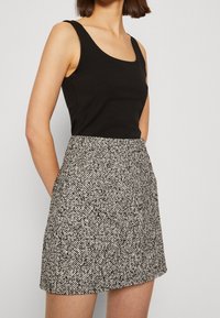 Žena v černém bezramínkovém topu s kulatým výstřihem a černobílé texturované minisukni vysokého pasu, s rukama v kapsách.