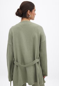 Cardigan lungo a maniche lunghe verde oliva in morbido tessuto testurizzato, con cintura annodata in vita e vestibilità rilassata e ampia.