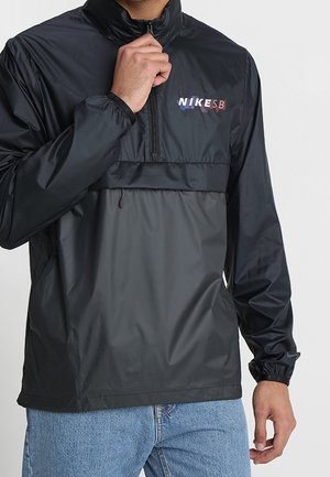 Homme portant une veste coupe-vent Nike SB noire avec demi-fermeture éclair, poignets élastiques et jean bleu clair, ajustant la fermeture éclair du col avec la main.