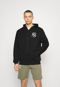 Sudadera negra con cremallera y un logo "NY" en el pecho izquierdo, bolsillos frontales y capucha con cordón. Combinada con pantalones cortos verde oliva.