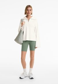 Pullover bianco in pile con colletto con zip, pantaloni corti da ciclismo verdi e sneakers bianche. Tieni una borsa imbottita grigio chiaro. Sfondo neutro.